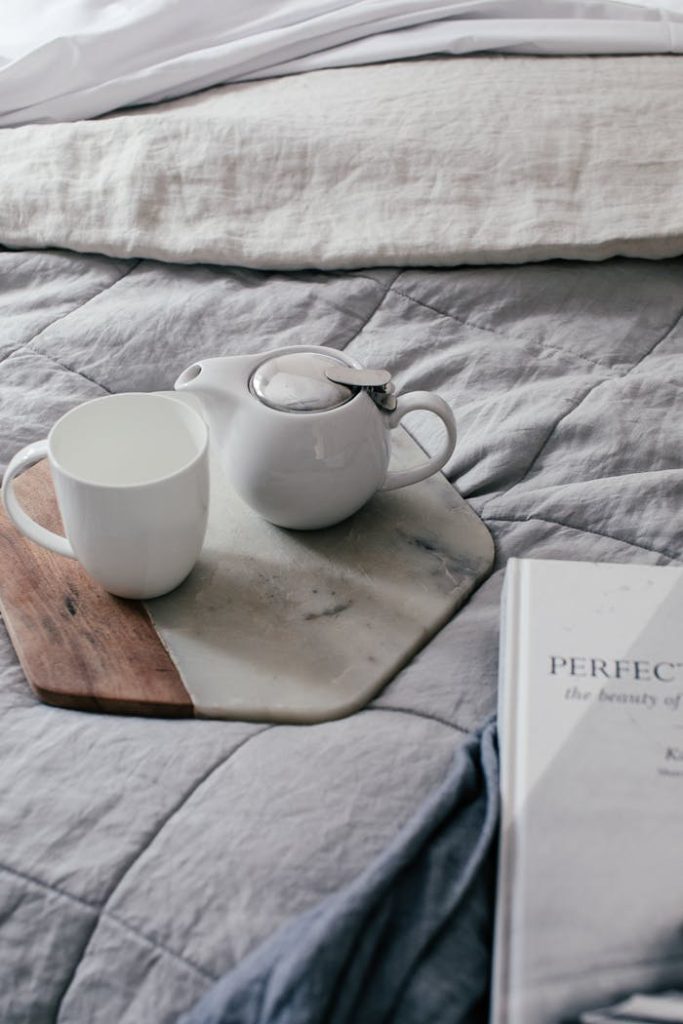 From above of mug near small teapot on marble and wooden tray on soft bed in hotel room